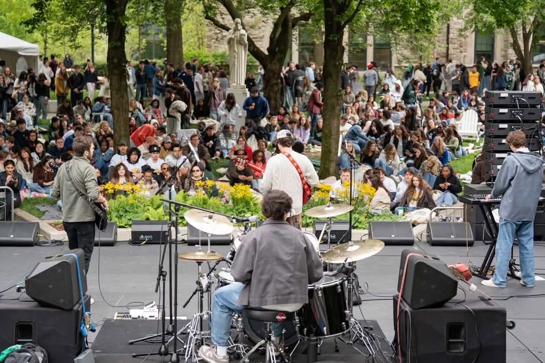 The view from behind the band, looking out at a large crowd seated on the grass at an outdoor campus show